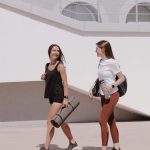 Two women in active wear walking outdoors while smiling and carrying yoga mats.