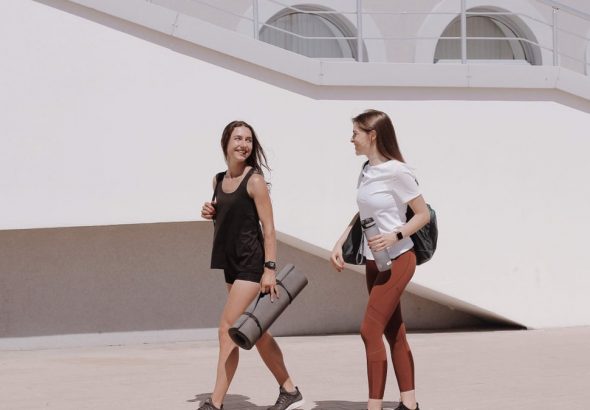 Two women in active wear walking outdoors while smiling and carrying yoga mats.